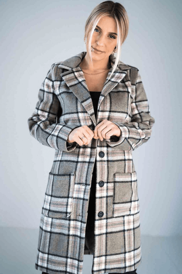 A beige plaid longline coat with a classic button-up design, large front pockets, and a tailored fit, worn by a woman with blonde hair styled in a low bun.






