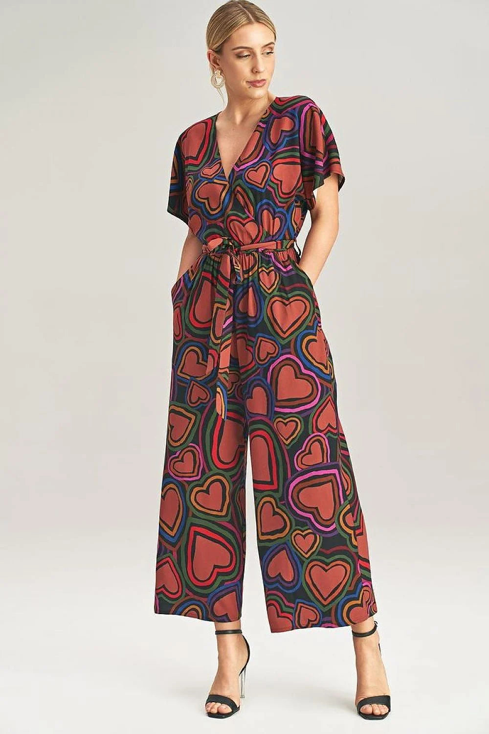 A woman wearing a wide-leg jumpsuit featuring a bold heart pattern, short sleeves, a V-neckline, and a tie belt at the waist.